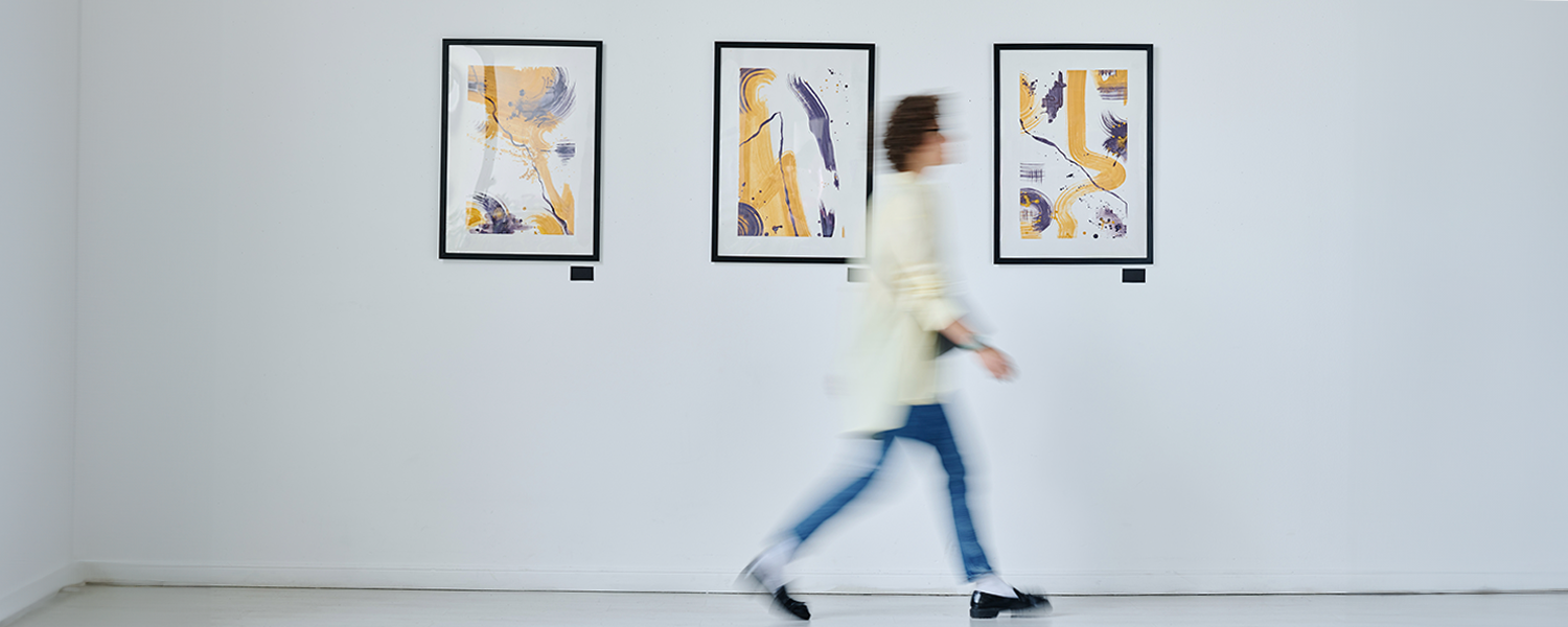 Abstract art exhibition featuring three framed artworks in yellow and purple, with a person walking past the display in a minimalistic gallery.