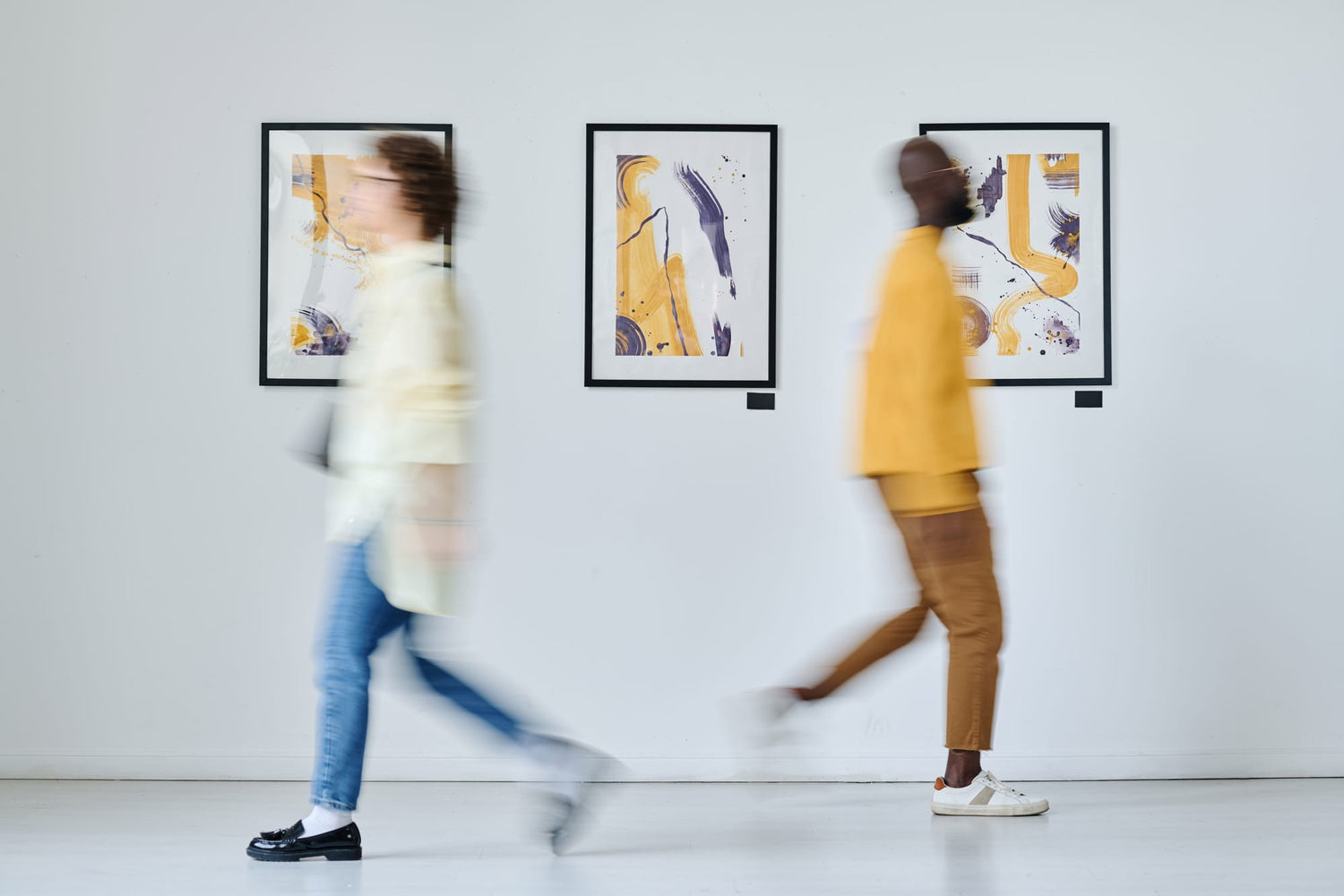 People walking past a gallery wall displaying three abstract wall art prints with yellow and purple accents in a modern art gallery.