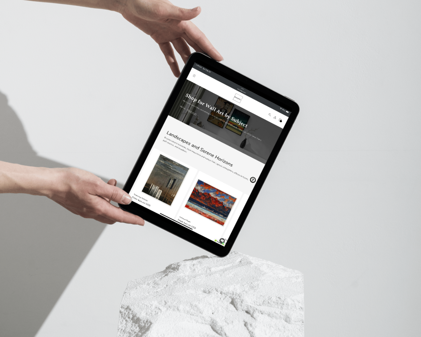 Hands holding a tablet displaying the Hive Artes website, featuring landscape wall art prints like 'Towering Solitude' and 'Crimson Peaks' on a minimalistic background.