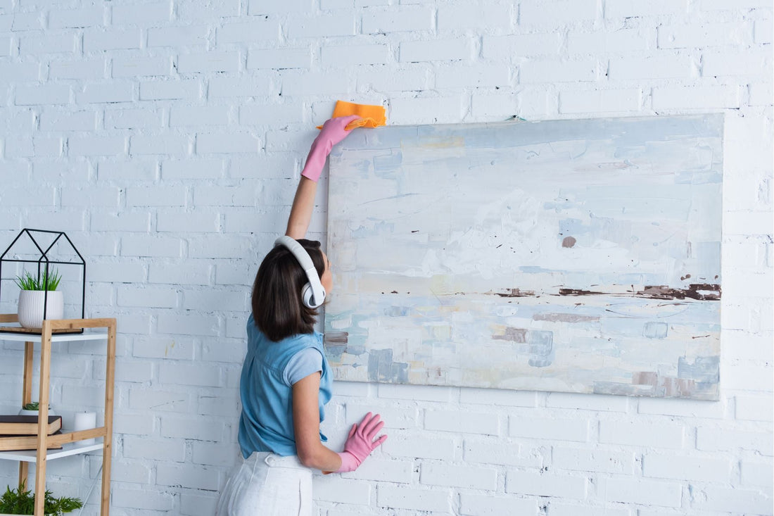 Woman dusting large canvas wall art during spring cleaning, wiping the top of the artwork with a cloth