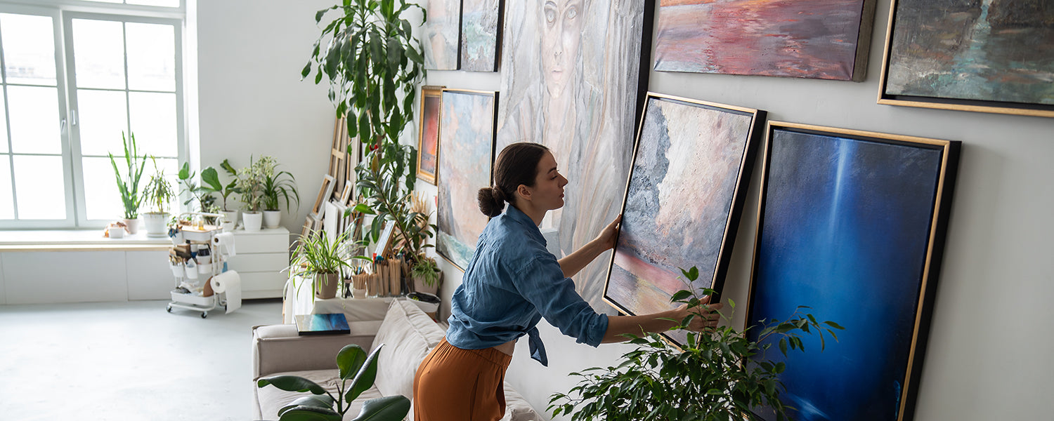 Woman hanging a framed canvas painting on a gallery wall filled with colorful fine art in a bright studio.