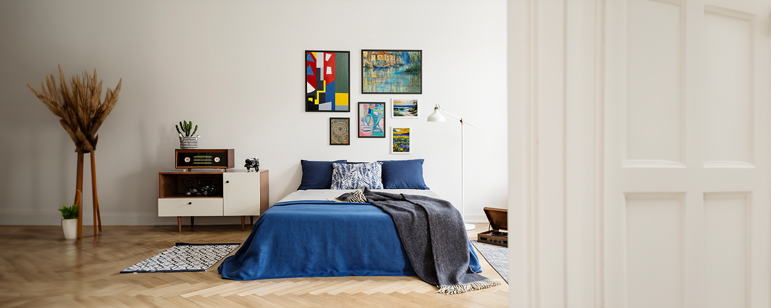 Cozy bedroom with blue bedding, a mid-century modern nightstand, and a gallery wall featuring abstract and landscape paintings in a variety of styles.