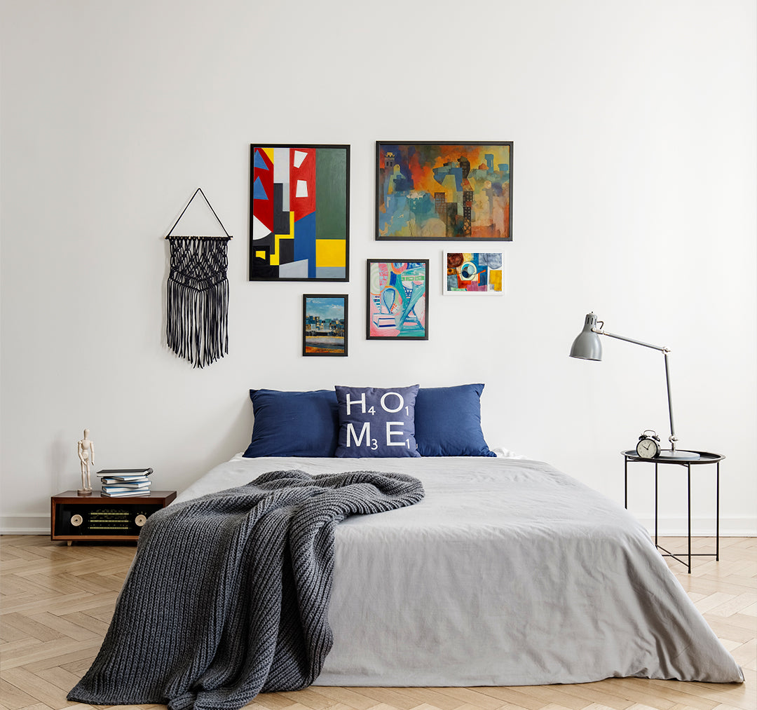 Modern bedroom with gray bedding, blue pillows, and a gallery wall of colorful abstract and geometric artworks, paired with a black macrame wall hanging.